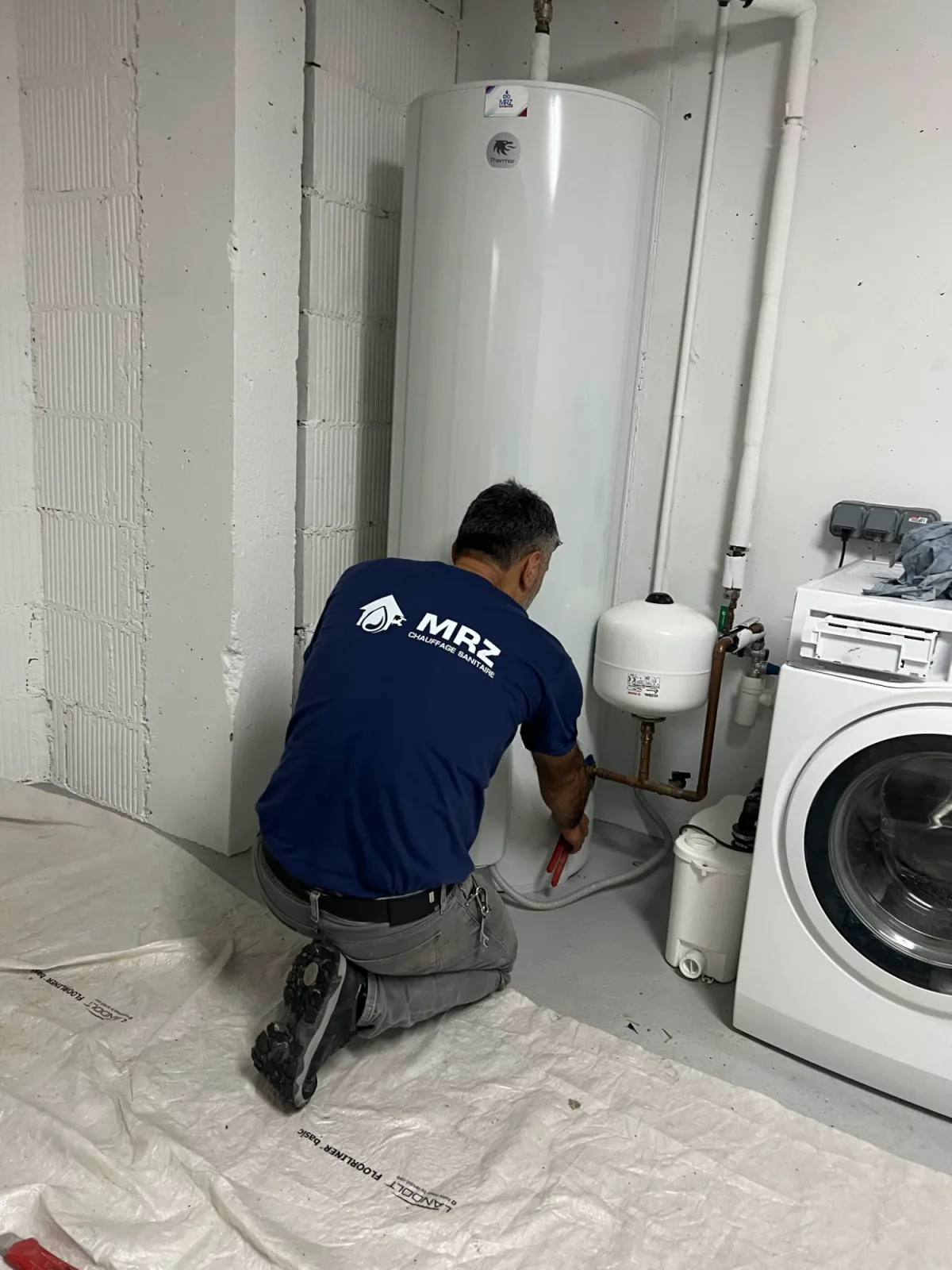 L'artisan de PLOMBERIE SANITAIRE MRZ en intervention sur un chauffe-eau, sol protégé par une bâche